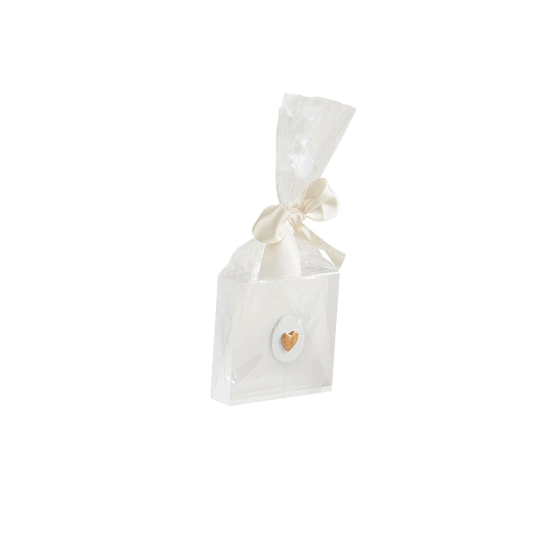 Studio photograph of an elegant white favor box featuring a small metallic gold heart on a textured oval plaque, wrapped in clear cellophane and tied with a cream satin ribbon bow against a white background