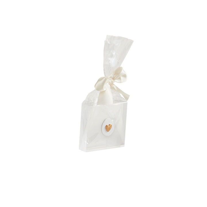 Studio photograph of an elegant white favor box featuring a small metallic gold heart on a textured oval plaque, wrapped in clear cellophane and tied with a cream satin ribbon bow against a white background