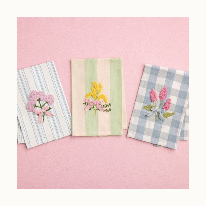 Embroidered linen tea towels featuring three floral designs on a pink textured background, including a blue-striped cloth with pink hydrangeas, a green-striped cloth with yellow mimosas, and a blue gingham cloth with pink blossoms and decorative velvet bows