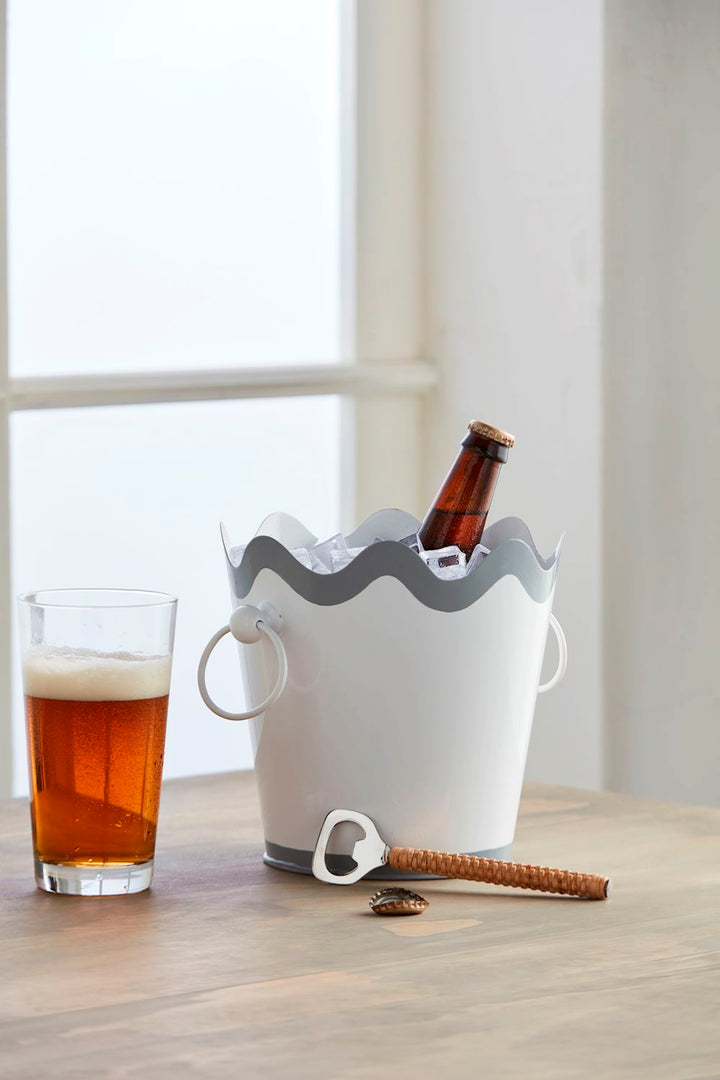 Photograph of a white ice bucket with a grey scalloped rim and ring handles on a wooden table featuring a tall glass of frosty amber beer and a bottle opener with a woven wicker handle set against a bright window background