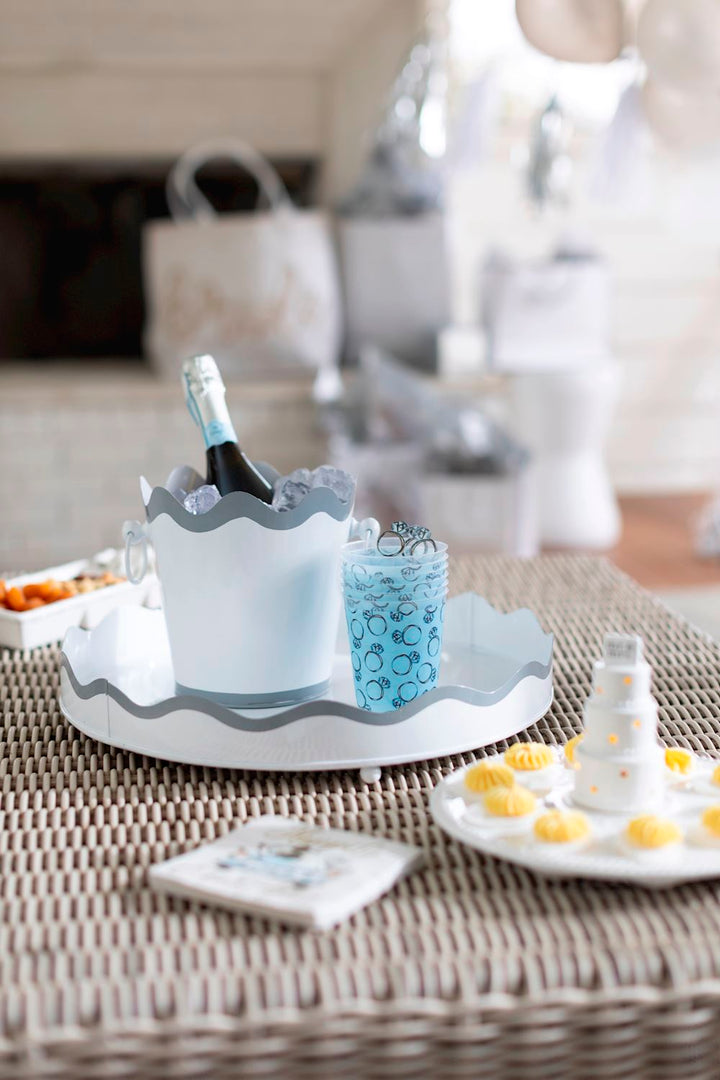 Photograph of a bridal shower refreshment station on a wicker table featuring a white scalloped ice bucket with chilled champagne, light blue cups with engagement ring patterns, and a miniature three-tier cake surrounded by yellow floral-topped treats