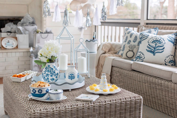 Photograph of an elegant engagement party celebration on a wicker-furnished sunroom porch featuring blue and white chinoiserie pottery, a decorative pillow with a diamond ring icon, and a miniature white tiered cake figurine