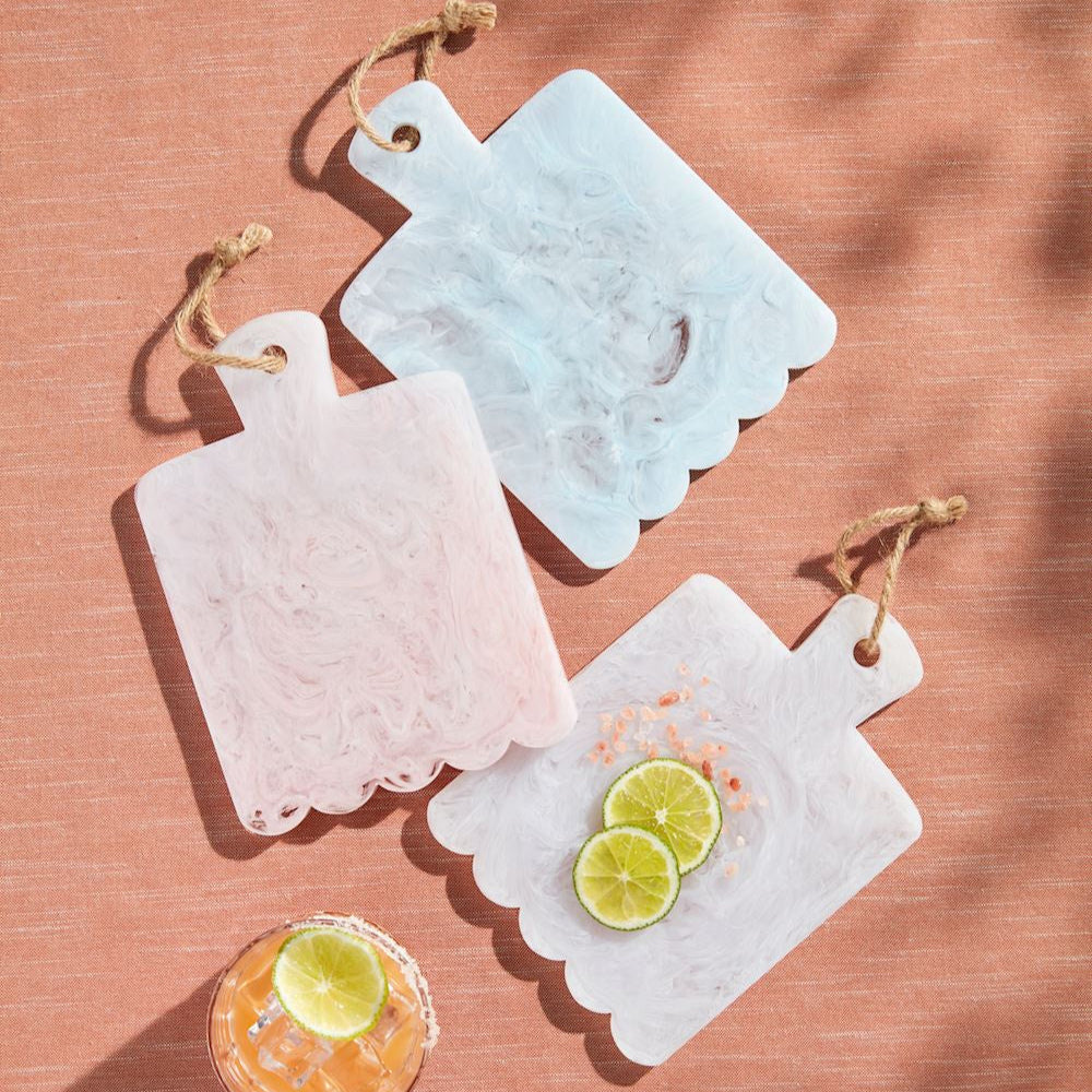Resin marble-effect scalloped mini serving boards in pastel pink, blue, and white are presented on a textured terracotta surface featuring rustic jute twine loops, vibrant lime slices, and coarse pink sea salt crystals