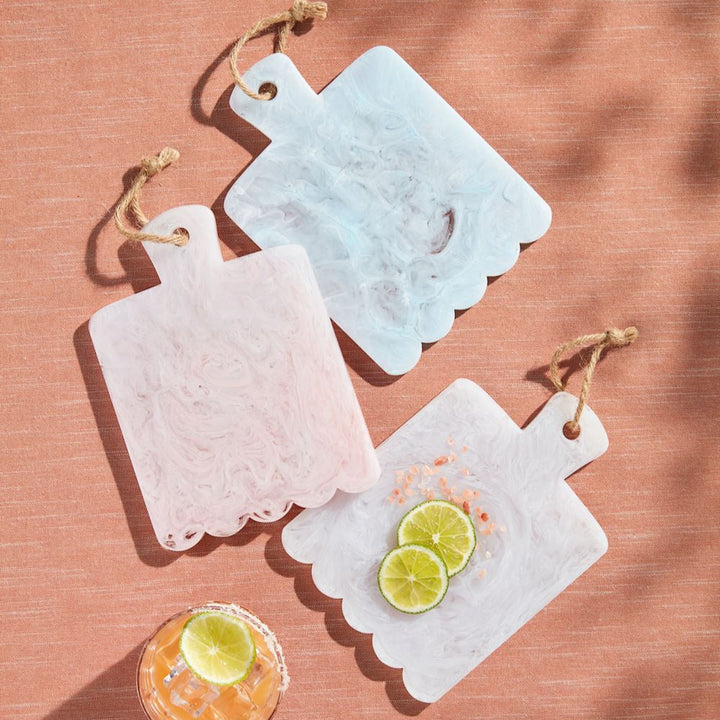 Resin marble-effect scalloped mini serving boards in pastel pink, blue, and white are presented on a textured terracotta surface featuring rustic jute twine loops, vibrant lime slices, and coarse pink sea salt crystals