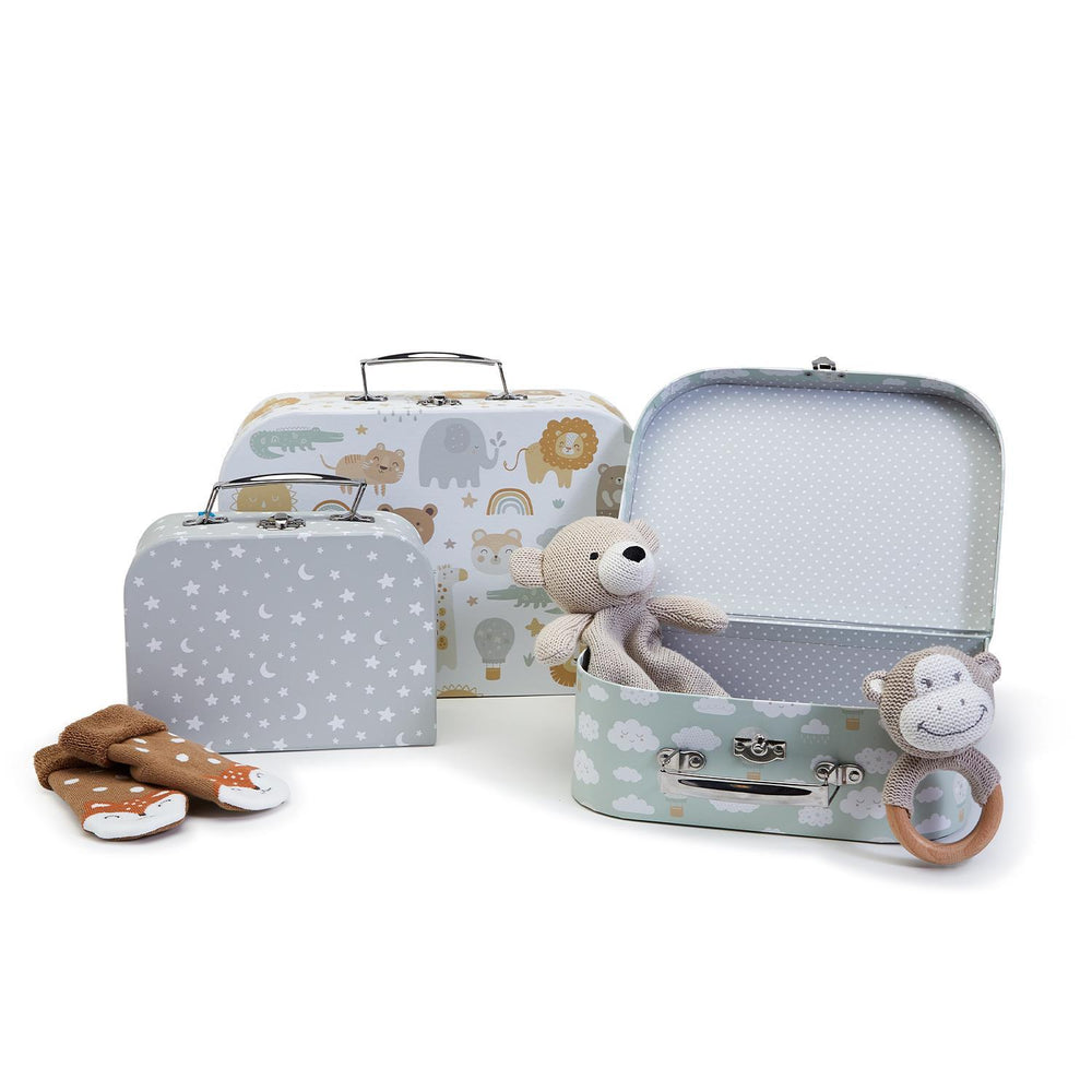 Product photograph of three decorative children's nesting suitcases with safari, celestial, and cloud patterns, featuring a beige knitted bear, a monkey wooden ring rattle, and fox-patterned booties on a white background