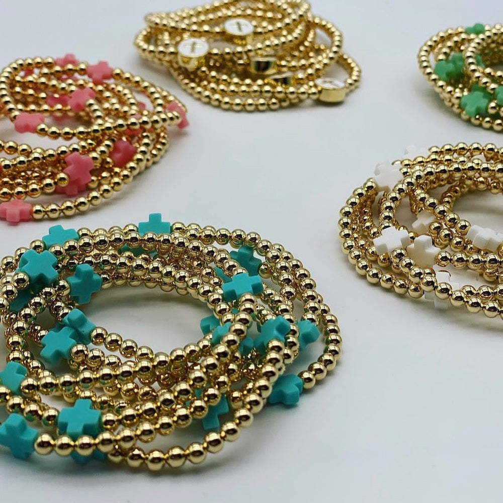 Jewelry photograph of multiple stacks of gold bead bracelets featuring vibrant teal, pink, and white cross-shaped charms arranged on a clean white surface