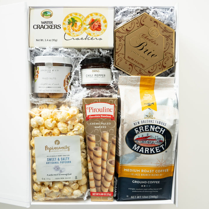 Still life photograph of a gourmet food gift box featuring New Orleans’ French Market Coffee and Popinsanity popcorn nestled in white crinkle paper with a gold hexagonal cheese box and chocolate hazelnut wafers