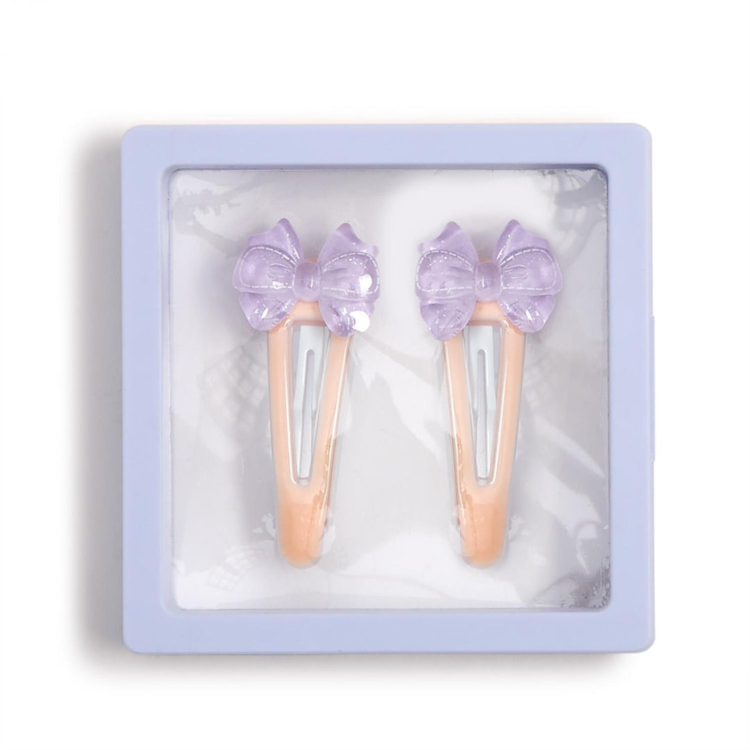 Product photograph of two peach-colored snap hair clips with lavender translucent bows featuring embossed stitch patterns displayed inside a light purple square frame with a clear window