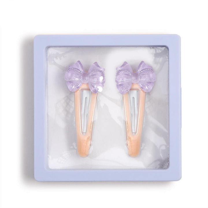 Product photograph of two peach-colored snap hair clips with lavender translucent bows featuring embossed stitch patterns displayed inside a light purple square frame with a clear window