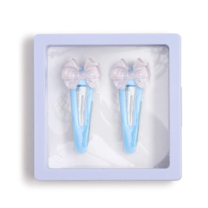 Product photography of a pair of light blue snap barrettes featuring translucent iridescent bows and ribbon-shaped accents displayed within a square lilac floating frame against a white background