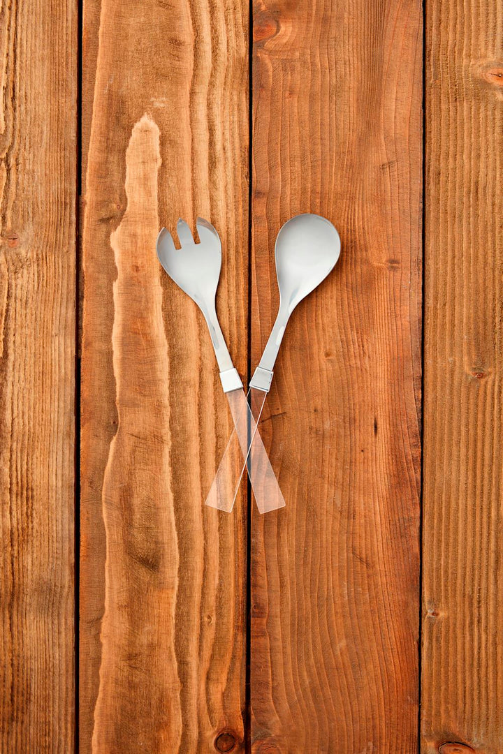 Digital photograph of a modern salad serving set on a rustic wooden surface featuring polished stainless steel heads and transparent handles arranged over textured vertical planks with rich grain and warm brown tones