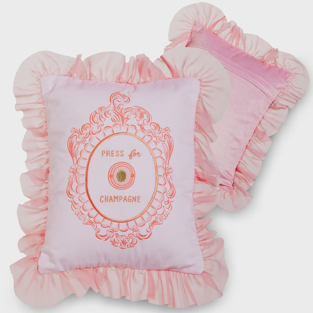 Decorative embroidered fabric pillows displayed on a neutral studio background featuring a pink ruffled border, an ornate coral filigree frame with the text PRESS for CHAMPAGNE, and a shimmering gold glitter button