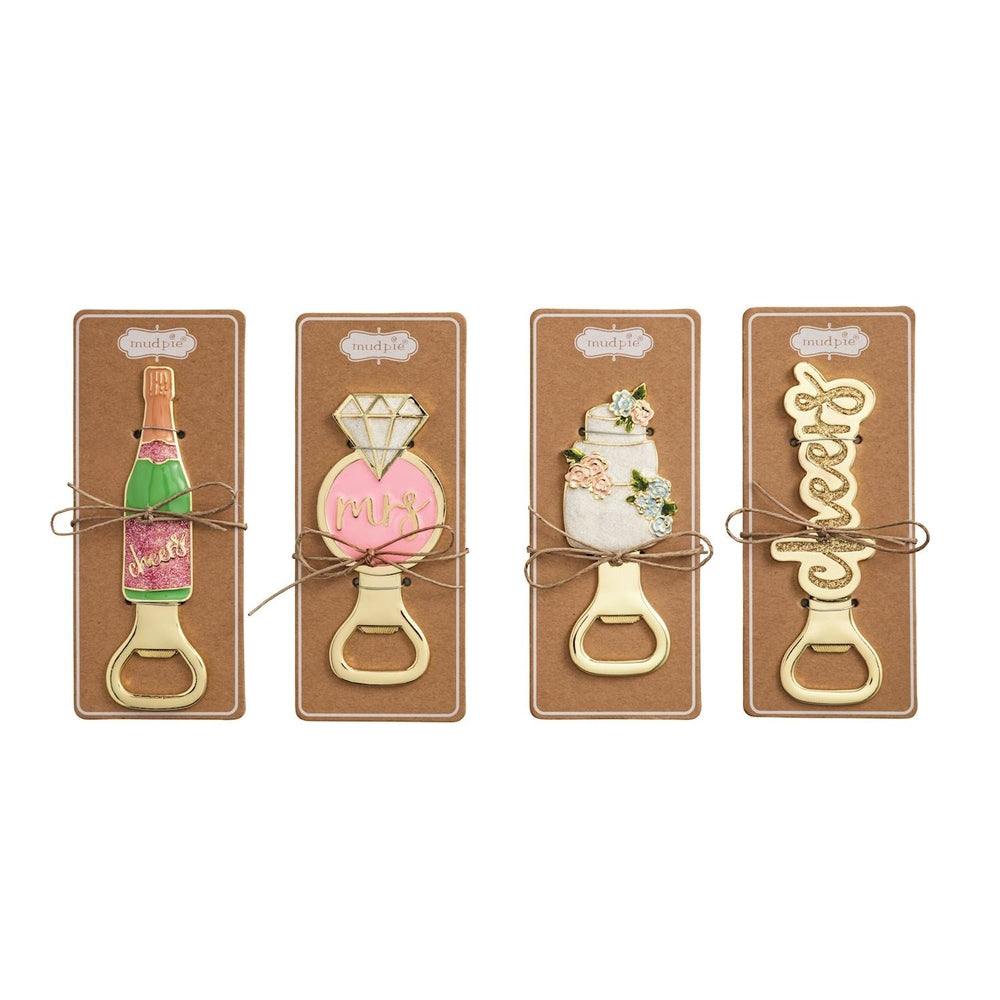 Enamel and gold-tone metal Mud Pie bottle openers displayed on kraft paper cards featuring a pink glitter champagne bottle, a floral-accented tiered cake, and a diamond ring tied with rustic twine