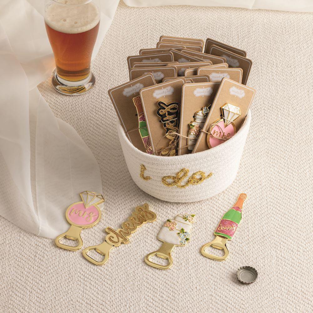 Commercial photography for Mud Pie features gold wedding-themed bottle openers including a pink engagement ring and a glittery green champagne bottle displayed in a white rope basket with gold I do embroidery on a textured surface beside a glass of beer