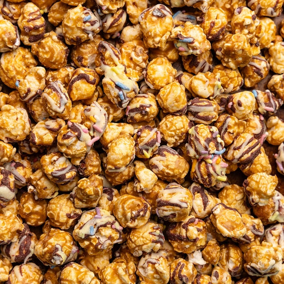 Digital macro photography of gourmet caramel popcorn in a dense pile featuring dark and white chocolate drizzles and colorful pastel sprinkles