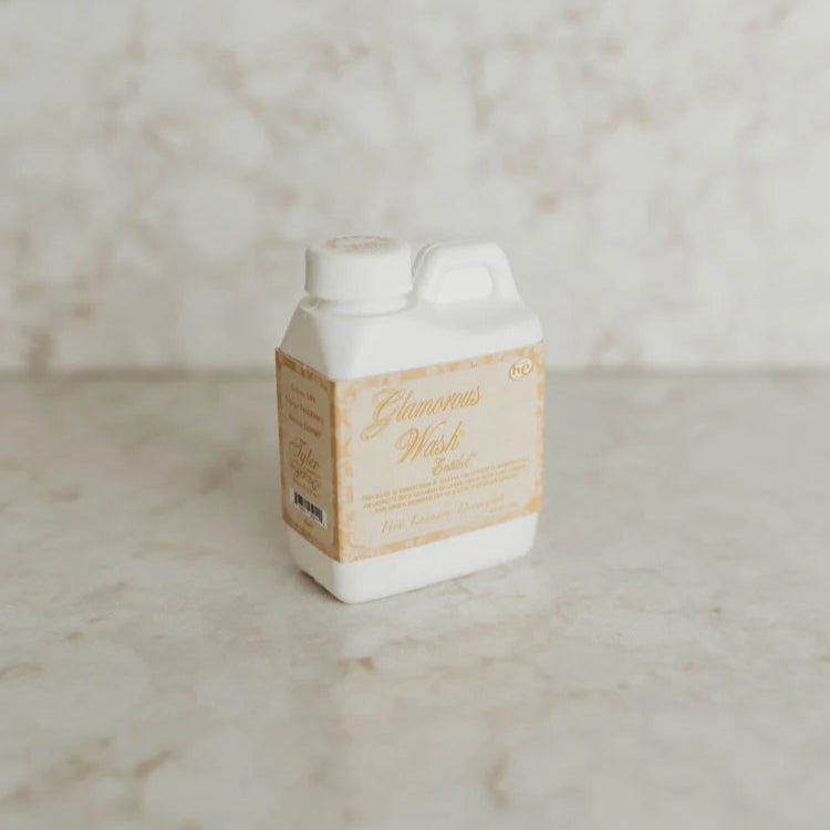 Photograph of a Tyler Candle Company Glamorous Wash laundry detergent jug on a beige marble countertop featuring a decorative gold and cream label with elegant cursive script and a white plastic handle