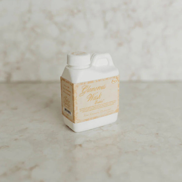 Photograph of a Tyler Candle Company Glamorous Wash laundry detergent jug on a beige marble countertop featuring a decorative gold and cream label with elegant cursive script and a white plastic handle
