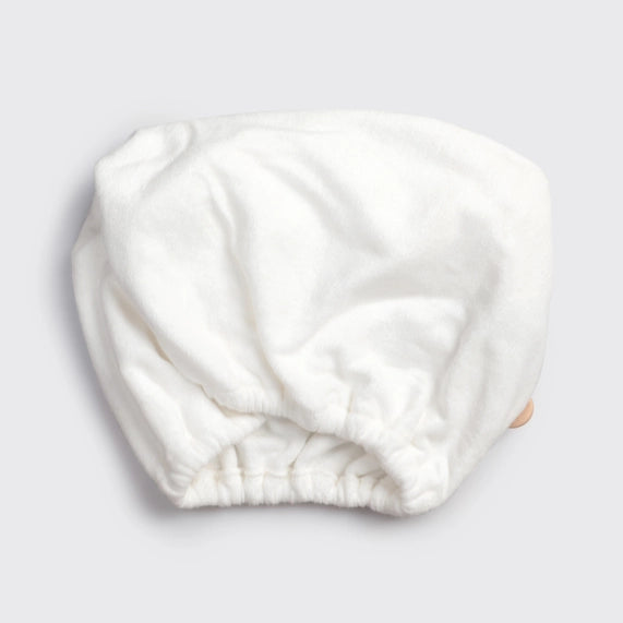 Studio product photograph of a white microfiber hair wrap on a neutral grey background featuring a gathered elasticated opening and a soft plush texture with a small tan button detail