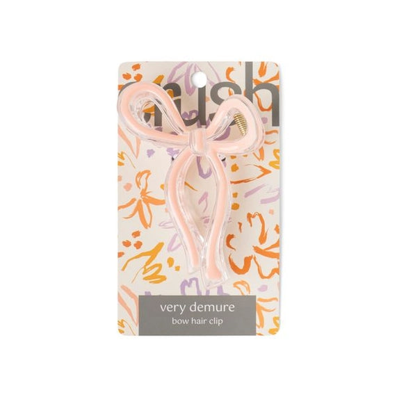 Product photograph of a Shush very demure bow hair clip featuring a pale pink translucent acrylic ribbon design and gold-toned spring mounted on an orange and lavender floral display card