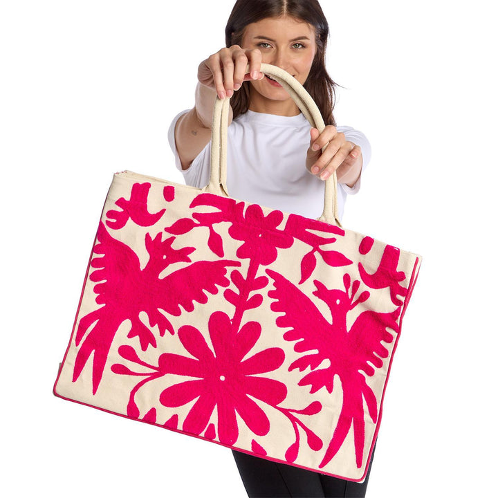 Hand-embroidered Tenango-style Otomi art on a large cream canvas tote bag held by a woman in a studio setting, showcasing vibrant fuchsia bird motifs, intricate floral patterns, and sturdy woven rope handles