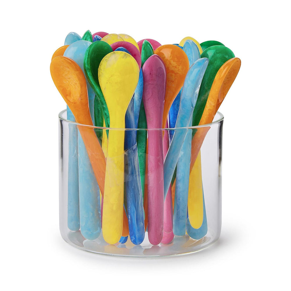 Resin spoons by Dinosaur Designs are gathered in a clear glass cylinder, showcasing vibrant marbled textures and a colorful palette of marigold yellow, hot pink, and sky blue with a smooth polished finish