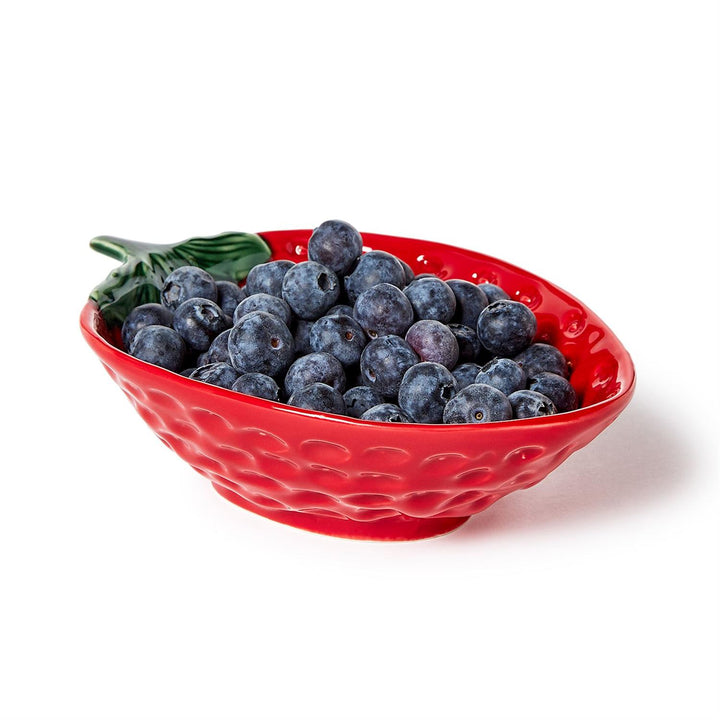 Ceramic strawberry-shaped bowl brimming with fresh blueberries on a white studio background, featuring a vibrant red dimpled exterior and a realistic green leaf handle alongside fruit with a dusty blue bloom