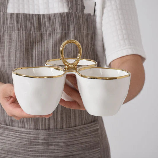 Ceramic triple serving bowl held by a person in a striped apron featuring scalloped gold-rimmed edges and a central polished gold ring handle