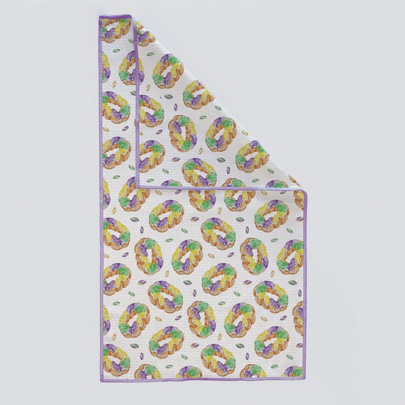 Watercolor print on a waffle-weave kitchen towel featuring a repeating pattern of iconic New Orleans Mardi Gras King Cakes decorated with vibrant purple, green, and gold sugar sprinkles and a thin purple border