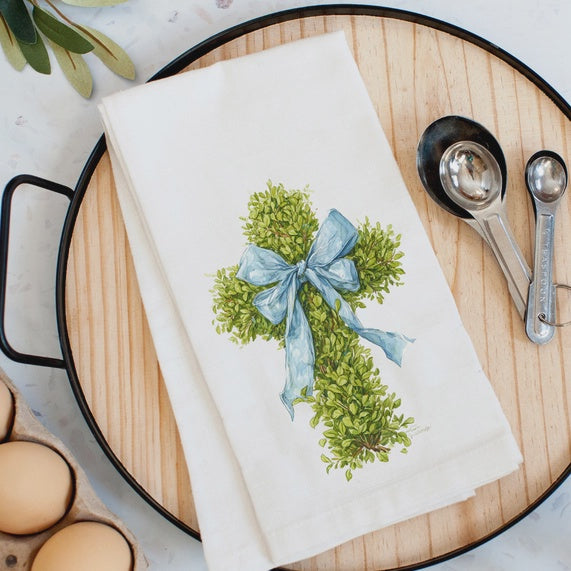 Watercolor illustration of a green boxwood cross with a soft blue ribbon bow on a white linen tea towel displayed on a round wooden tray next to stainless steel measuring spoons and fresh brown eggs