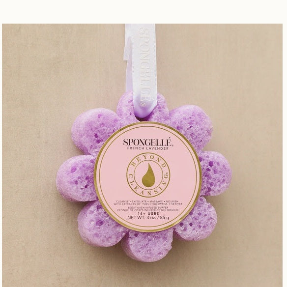 Product photograph by Spongellé of a purple flower-shaped French Lavender body wash infused buffer featuring a porous lavender texture, a circular pink label with gold foil accents, and a white hanging ribbon against a neutral beige background