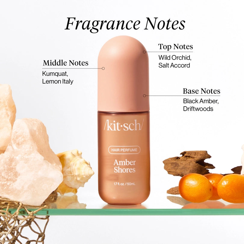 Commercial product photography for Kitsch features the Amber Shores hair perfume bottle on a glass shelf surrounded by textured pink salt rocks, a spiraled seashell, and bright orange kumquats illustrating the various fragrance notes