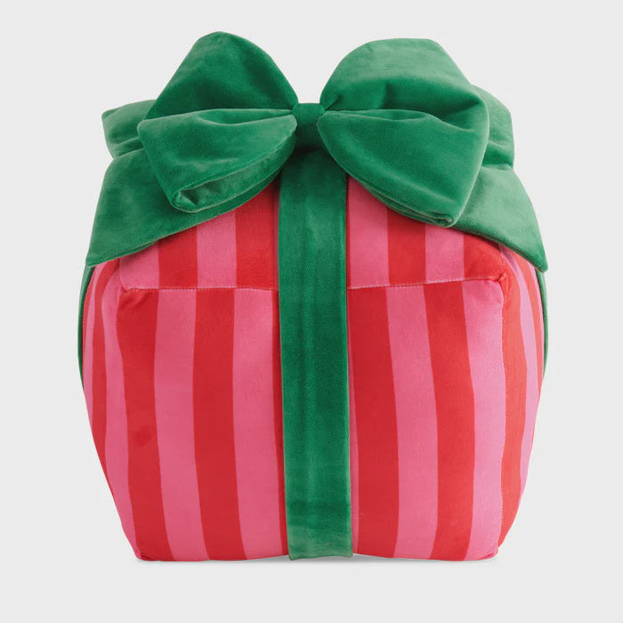 Plush velvet decorative pillow shaped like a gift box in a neutral studio setting featuring bold red and pink vertical stripes and an oversized emerald green bow tied on top