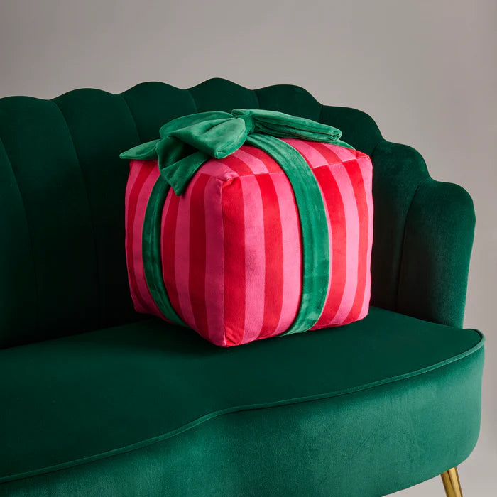 Velvet plush decorative pillow shaped like a wrapped gift resting on a dark green scalloped sofa, featuring vibrant pink and red vertical stripes and an oversized green velvet bow on top