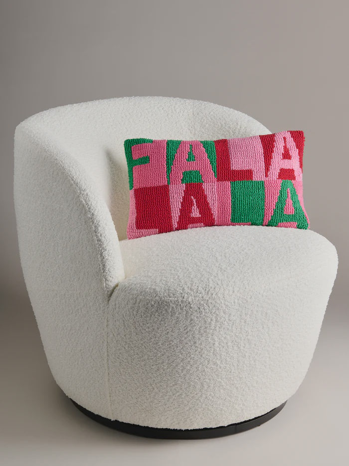 Product photograph of a white bouclé swivel armchair featuring a festive rectangular hook-stitch pillow with FA LA LA LA lettering in a red pink and green checkerboard pattern set against a minimalist studio background