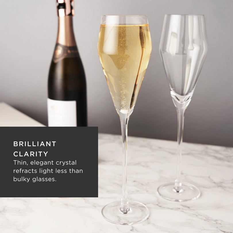 Commercial product photograph of two elegant crystal champagne flutes on a white marble surface featuring one glass filled with bubbly golden liquid and a champagne bottle with rose gold foil in the background