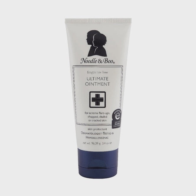 Product photography of a Noodle & Boo Ultimate Ointment tube features the brand's signature profile silhouettes, a black medical cross icon, and a dark navy blue cap set against a plain gray studio backdrop