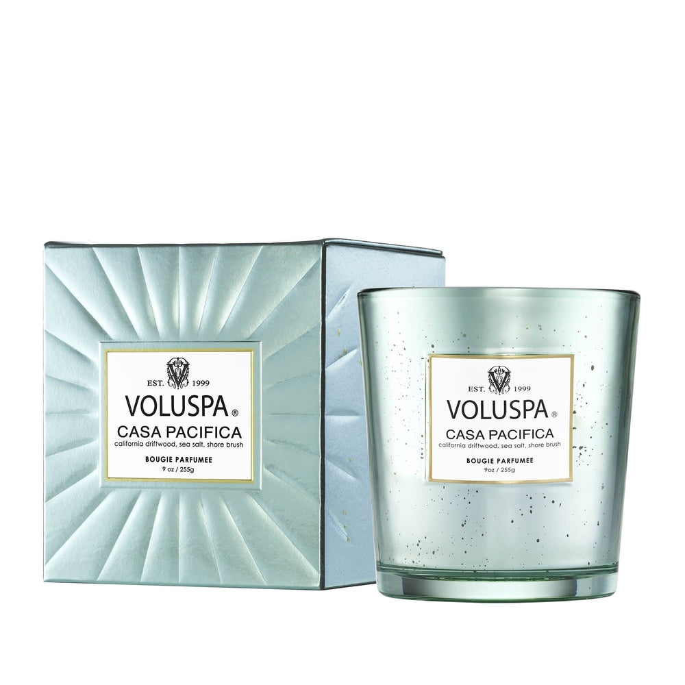 Product photography of a Voluspa Casa Pacifica scented candle and its matching box featuring an embossed sunburst pattern, a speckled seafoam glass vessel, and gold-accented labels on a white backdrop
