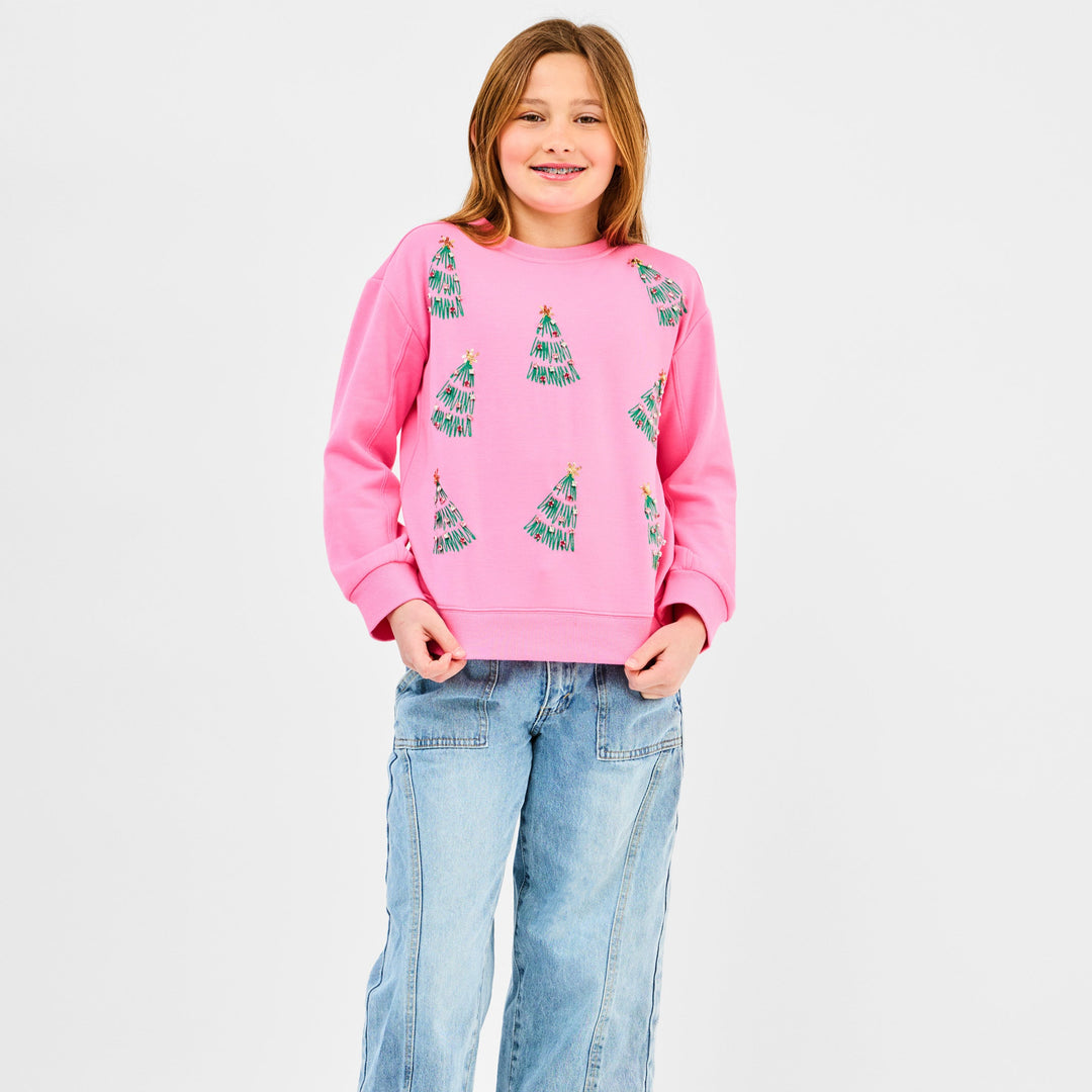 Studio photograph of a young girl in a white studio wearing a pink holiday sweatshirt with green sequined Christmas trees and light-wash jeans while smiling with dental braces
