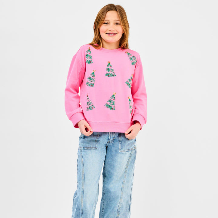 Studio photograph of a young girl in a white studio wearing a pink holiday sweatshirt with green sequined Christmas trees and light-wash jeans while smiling with dental braces