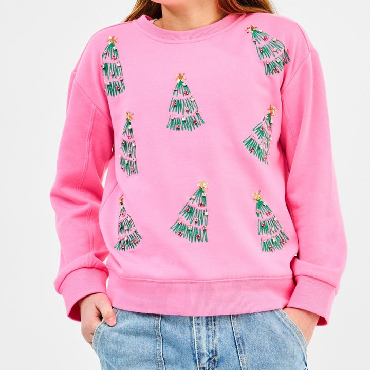 Studio photograph of a model wearing a bubblegum pink sweatshirt decorated with green embroidered Christmas trees featuring gold star sequins and colorful bead ornaments against a white background