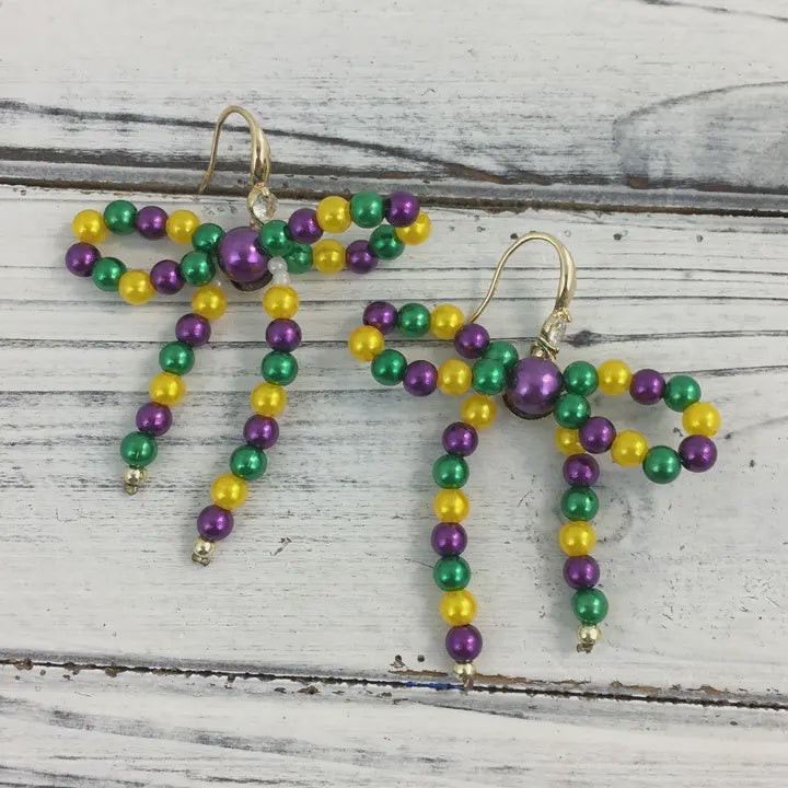 Handcrafted beaded jewelry featuring Mardi Gras themed bow earrings with metallic purple green and gold beads and gold-tone French hooks displayed on a rustic white wooden background