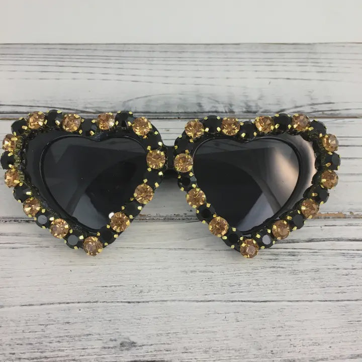 Handcrafted embellished eyewear featuring black heart-shaped frames adorned with amber and onyx rhinestones resting on a weathered white wooden surface