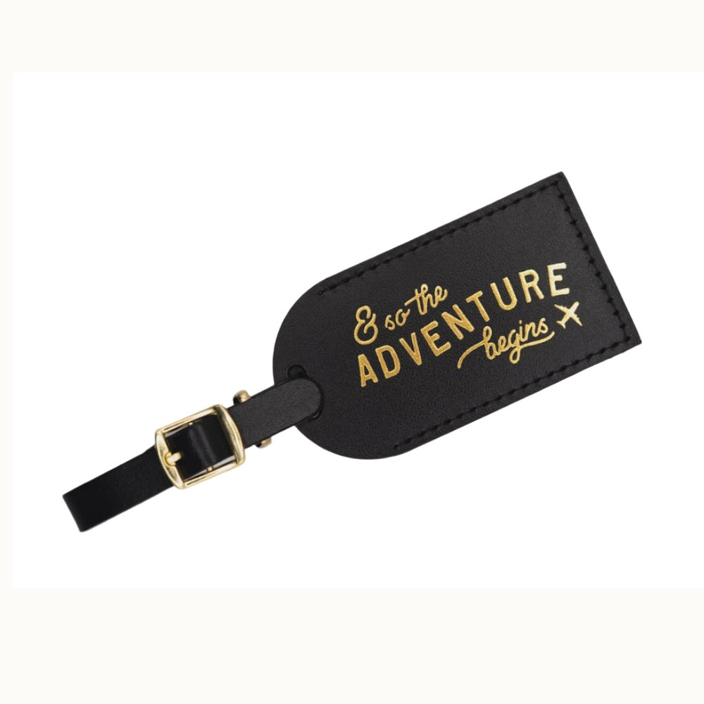Black leather luggage tag on a white background featuring gold foil typography reading and so the adventure begins, a small airplane icon, and a polished gold buckle with perimeter stitching