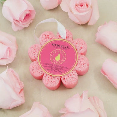 Product photography by Spongellé of a pink flower-shaped Bulgarian Rose body buffer featuring a white hanging ribbon and textured surface surrounded by soft pink rosebuds on a neutral background