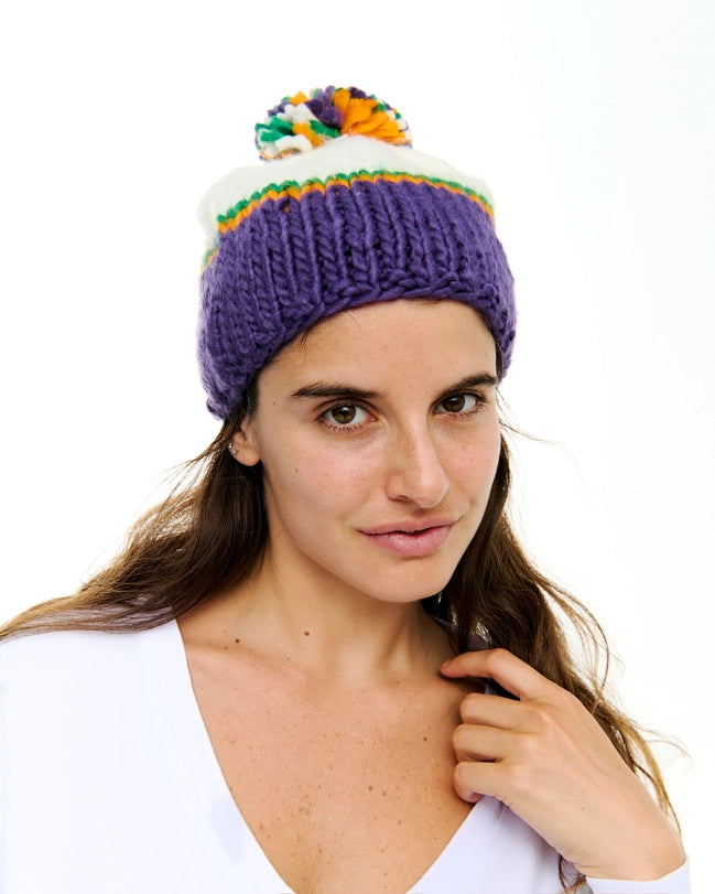 Studio photograph of a woman wearing a colorful knit beanie against a white background featuring a chunky purple brim, thin orange and green stripes, and a multicolored pom-pom