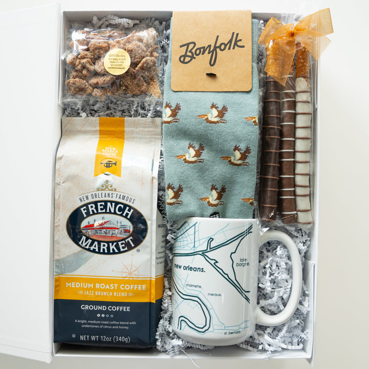 Photograph of a New Orleans themed gift box containing a bag of French Market coffee, a white ceramic mug with a city map, teal pelican patterned socks, and chocolate-covered pretzels with a translucent orange bow resting on white crinkle paper