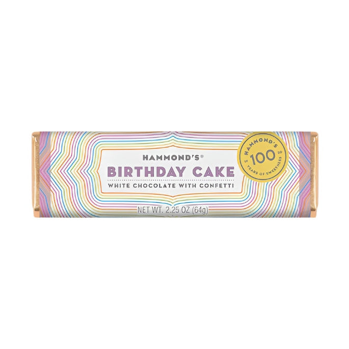 Product photography by Hammond's of a Birthday Cake white chocolate bar featuring a vibrant multi-colored zig-zag pattern, a gold 100-year anniversary seal, and bold purple typography on a white studio background