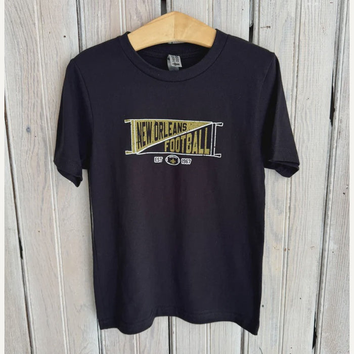 Screen-printed black cotton t-shirt featuring a New Orleans football graphic on a wooden hanger against a white plank wall with a distressed gold-toned pennant design and iconic fleur-de-lis symbol