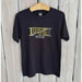 Screen-printed black cotton t-shirt featuring a New Orleans football graphic on a wooden hanger against a white plank wall with a distressed gold-toned pennant design and iconic fleur-de-lis symbol
