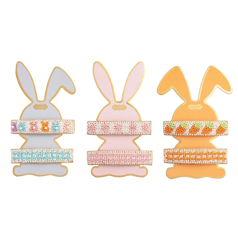 Handcrafted beaded hair clips by Mud Pie are displayed on blue, pink, and orange bunny-shaped cardstock holders on a white background featuring pastel seed beads arranged in carrot, bunny, and floral patterns with gold-trimmed edges